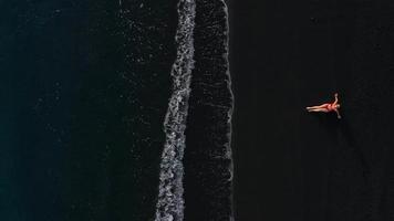 parte superior ver de un niña en un rojo traje de baño acostado en un negro playa en el navegar línea. costa de el isla de tenerife, canario video