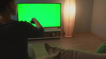 donna è seduta su il divano, Guardando tv con un' verde schermo, commutazione canali con un' a distanza controllo. indietro Visualizza. croma chiave. in casa video