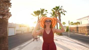 feliz mulher dentro uma grande amarelo chapéu caminhando ao longo uma Palma beco às pôr do sol video
