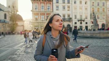 donna indossare un' grigio cappotto con un' thermos tazza nel mano a piedi giù un vecchio strada utilizzando smartphone a tramonto. comunicazione, sociale reti, in linea shopping concetto. video