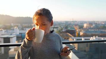 mujer empieza su día con un taza de té o café y comprobación correos electrónicos en su teléfono inteligente en el balcón a amanecer, lento movimiento. moderno urbano estilo de vida video