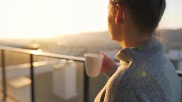 vrouw begint haar dag met een kop van thee of koffie en controle e-mails in haar smartphone Aan de balkon Bij ochtendgloren, langzaam beweging. modern stedelijk levensstijl video