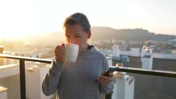 mujer empieza su día con un taza de té o café y comprobación correos electrónicos en su teléfono inteligente en el balcón a amanecer, lento movimiento. moderno urbano estilo de vida video