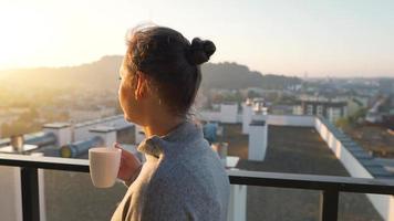 Woman starts her day with a cup of tea or coffee and checking emails in her smartphone on the balcony at dawn, slow motion. Modern urban lifestyle video
