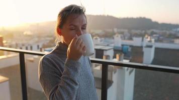 donna inizia sua giorno con un' tazza di tè o caffè e controllo messaggi di posta elettronica nel sua smartphone su il balcone a alba, lento movimento. moderno urbano stile di vita video