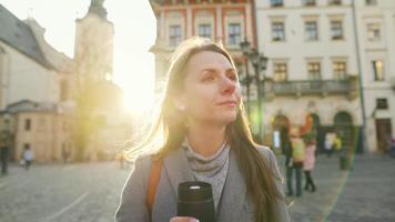 donna indossare un' grigio cappotto con un' thermos tazza nel mano a piedi giù un vecchio strada utilizzando smartphone a tramonto. comunicazione, sociale reti, in linea shopping concetto. video
