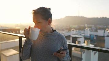 mujer empieza su día con un taza de té o café y comprobación correos electrónicos en su teléfono inteligente en el balcón a amanecer, lento movimiento. moderno urbano estilo de vida video