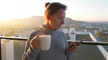 donna inizia sua giorno con un' tazza di tè o caffè e controllo messaggi di posta elettronica nel sua smartphone su il balcone a alba, lento movimento. moderno urbano stile di vita video