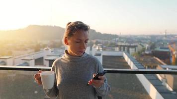 mujer empieza su día con un taza de té o café y comprobación correos electrónicos en su teléfono inteligente en el balcón a amanecer, lento movimiento. moderno urbano estilo de vida video