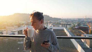 mujer empieza su día con un taza de té o café y comprobación correos electrónicos en su teléfono inteligente en el balcón a amanecer, lento movimiento. moderno urbano estilo de vida video