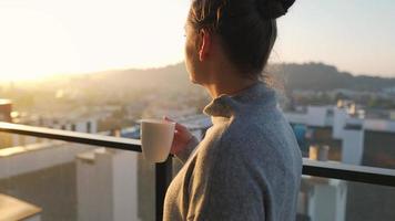 mujer empieza su día con un taza de té o café y comprobación correos electrónicos en su teléfono inteligente en el balcón a amanecer, lento movimiento. moderno urbano estilo de vida video