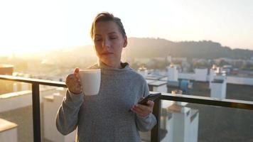 Woman starts her day with a cup of tea or coffee and checking emails in her smartphone on the balcony at dawn, slow motion. Modern urban lifestyle video