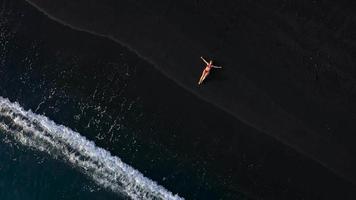 topo Visão do uma menina dentro uma vermelho roupa de banho deitado em uma Preto de praia em a surfar linha. costa do a ilha do tenerife, canário video