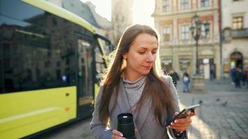Frau tragen ein grau Mantel mit ein Thermosflasche Tasse im Hand Gehen Nieder ein alt Straße mit Smartphone beim Sonnenuntergang. Kommunikation, Sozial Netzwerke, online Einkaufen Konzept. video