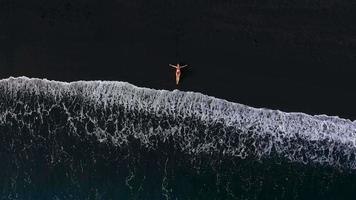 top visie van een meisje in een rood zwempak aan het liegen Aan een zwart strand Aan de surfen lijn. kust van de eiland van tenerife, kanarie video