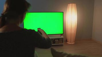 mujer es sentado en el sofá, acecho televisión con un verde pantalla, traspuesta canales con un remoto control. espalda vista. croma llave. adentro video