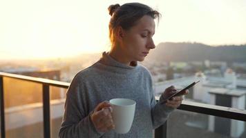 mujer empieza su día con un taza de té o café y comprobación correos electrónicos en su teléfono inteligente en el balcón a amanecer, lento movimiento. moderno urbano estilo de vida video