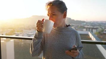 mulher começa dela dia com uma copo do chá ou café e verificação e-mails dentro dela Smartphone em a sacada às alvorecer, lento movimento. moderno urbano estilo de vida video