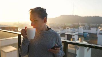 Woman starts her day with a cup of tea or coffee and checking emails in her smartphone on the balcony at dawn, slow motion. Modern urban lifestyle video