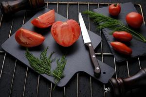 pizarra de tomates en rodajas, hojas de eneldo, dos molinillos de especias y un cuchillo con mango de madera en la parrilla. foto