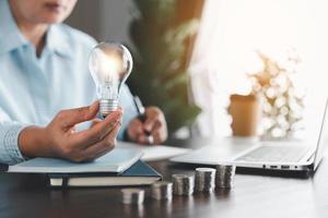 Businesswoman hand holding lightbulb with take notes on the table. Idea of saving energy and accounting finance in home and family, save world and energy power. Save energy plan concept. photo
