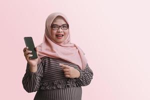 Portrait of cheerful Asian woman with hijab, showing blank screen mobile phone and pointing. isolated image on pink background photo