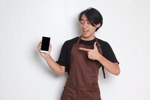 Portrait of attractive Asian barista man in brown apron showing blank screen of mobile phone. Empty space for mock up. Isolated on white background photo