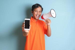 Portrait of attractive Asian man in orange shirt speaking louder using megaphone, promoting product while showing blank screen mobile phone. Advertising concept. Isolated image on blue background photo