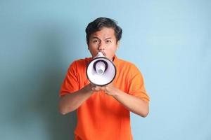 Portrait of attractive Asian man in orange shirt speaking louder using megaphone, promoting product. Advertising concept. Isolated image on blue background photo