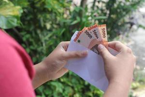 cerca arriba de masculino mano insertando cinco mil rupia dinero billete de banco dentro sobre. financiero y ahorros concepto foto