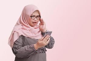 Portrait of shocked Asian woman with hijab screaming and gesturing in panic, while looking at mobile phone. Isolated image on pink background photo