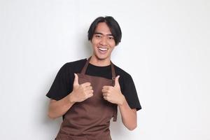 Portrait of attractive Asian barista man in brown apron showing thumb up hand gesture. Advertising concept. Isolated image on white background photo