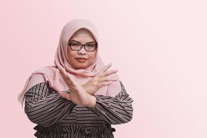 Portrait of serious Asian woman with hijab, showing X sign of hands for refusing an invitation from someone. Isolated image on pink background photo