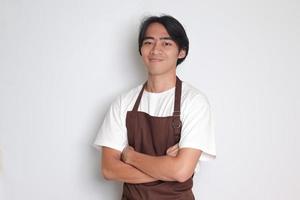 Portrait of attractive Asian barista man in brown apron keeping arms crossed and looking at camera. Advertising concept. Isolated image on white background photo