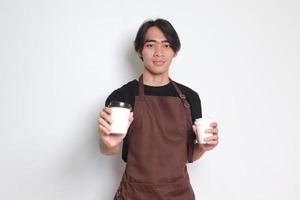 Portrait of attractive Asian barista man in brown apron holding two take away paper coffee cups. Isolated image on white background photo