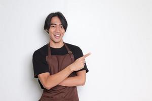 Portrait of attractive Asian barista man in brown apron showing product, pointing at something with hands. Advertising concept. Isolated image on white background photo