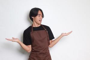 Portrait of attractive Asian barista man in brown apron choosing between 2 different options, holding two hands with empty space. Decision and choice concept. Isolated image on white background photo