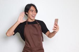 Portrait of attractive Asian barista man in brown apron making hi gesture while having conversation on video call. Isolated image on white background photo