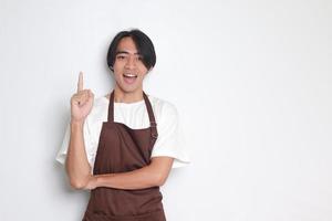 Portrait of attractive Asian barista man in brown apron showing product, pointing at something with hands. Advertising concept. Isolated image on white background photo