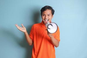 Portrait of attractive Asian man in orange shirt speaking louder using megaphone, promoting product. Advertising concept. Isolated image on blue background photo