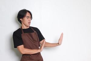 Portrait of attractive Asian barista man in brown apron forming a hand gesture to avoid something. Isolated image on white background photo