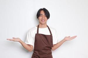 Portrait of attractive Asian barista man in brown apron choosing between 2 different options, holding two hands with empty space. Decision and choice concept. Isolated image on white background photo