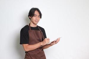 Portrait of attractive Asian barista man in brown apron taking order, writing on menu book list. Isolated image on white background photo