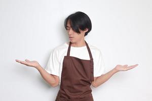 Portrait of attractive Asian barista man in brown apron choosing between 2 different options, holding two hands with empty space. Decision and choice concept. Isolated image on white background photo