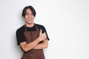 Portrait of attractive Asian barista man in brown apron showing thumb up hand gesture. Advertising concept. Isolated image on white background photo
