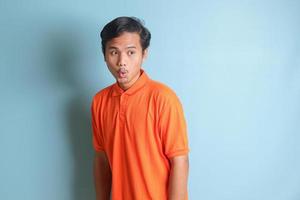 surprised Asian man in orange shirt standing against blue background, showing shocked expression and looking aside photo