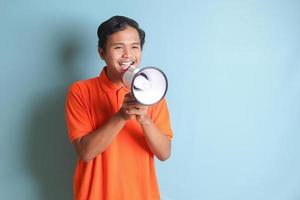 Portrait of attractive Asian man in orange shirt speaking louder using megaphone, promoting product. Advertising concept. Isolated image on blue background photo