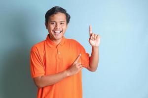 excited Asian man in orange shirt smiling and looking at the camera pointing with two hands and fingers to the side. Isolated image on blue background photo