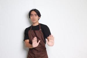 Portrait of attractive Asian barista man in brown apron forming a hand gesture to avoid something. Isolated image on white background photo