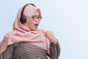 Portrait of cheerful Asian woman with hijab keeping hand near mouth as if microphone sings song along while listening to music using headphone. Isolated image on blue background photo